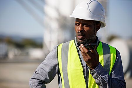 A construction worker wearing a hard hat and safety vest is talking on a walkie talkie.
